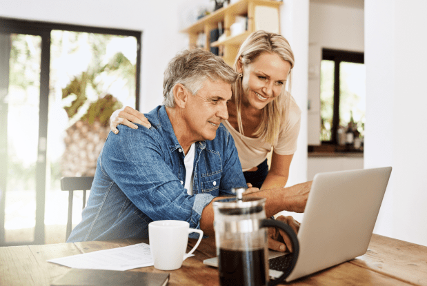 Older couple seated at a table in their home, looking at a laptop, representing financial planning services for retirement.